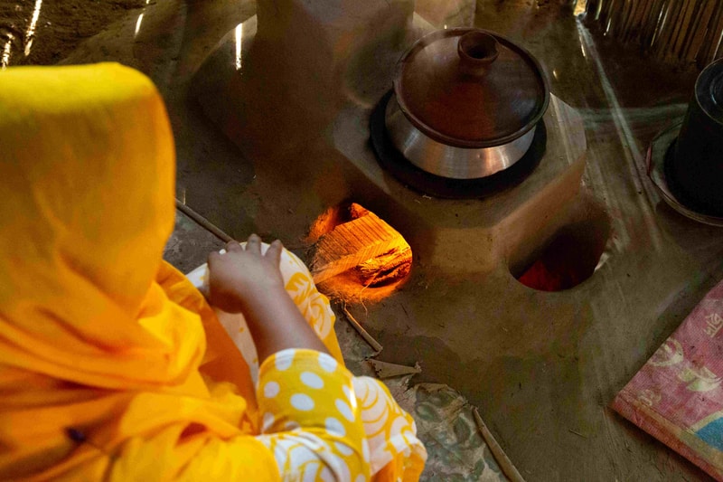 A person wearing a yellow robe or scarf is seated near a glowing wood fire in a traditional stove, tending to burning logs with a cloth nearby.