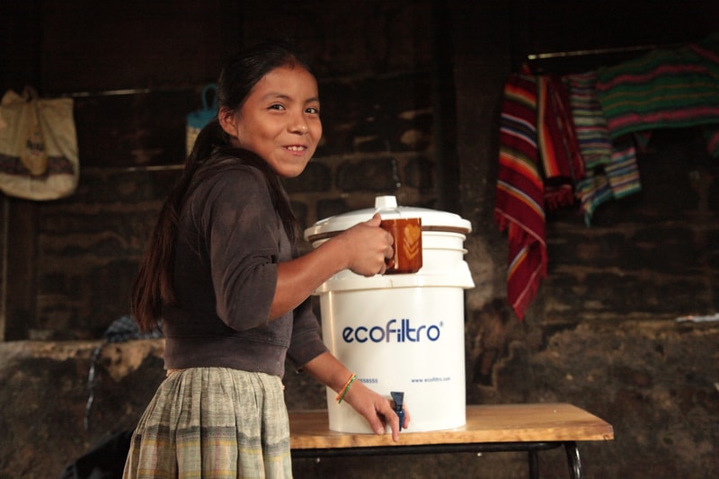A smiling woman with long dark hair holds a brown mug while standing beside a white Ecofiltro water filter on a wooden table inside a dim, rustic room with colorful blankets hanging in the background.