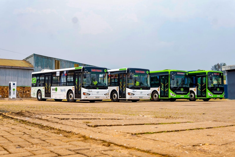 Multiple modern buses—three white with branding and two bright green—were parked in a line outside an industrial yard with a brick and metal building in the background.