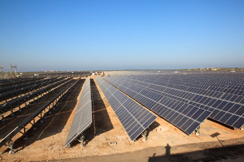 Rows of solar panels mounted on metal frames tilt along a desert-like landscape under a clear blue sky, forming a vast solar farm extending to the horizon.