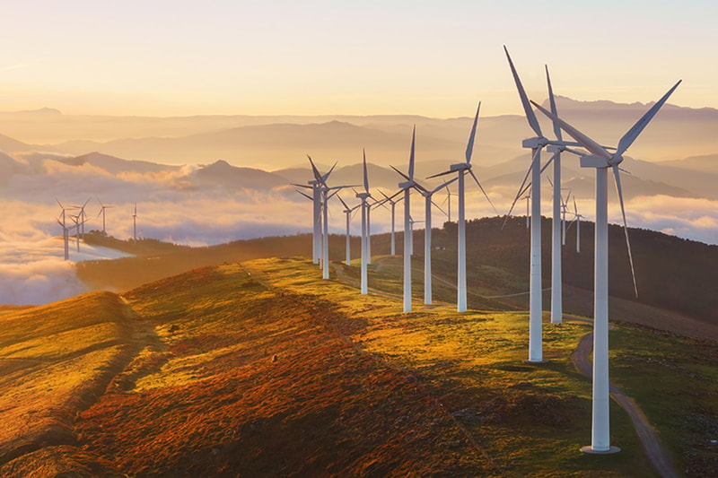 Wind turbines line a sunlit hilltop at golden hour, rows stretching into misty clouds over rolling mountains. clear sky, warm colors, scenic renewable energy landscape.