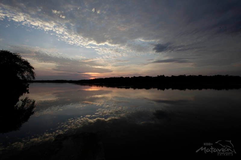 Sunset over a calm lake with a silhouetted treeline, the sky filled with soft clouds and warm hues mirrored on the water's surface, creating a tranquil reflection.