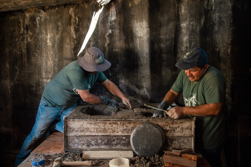 Two workers wearing green shirts and hats are shaping a dusty, clay-like material inside a large industrial mold, using tools at a dusty worktable.