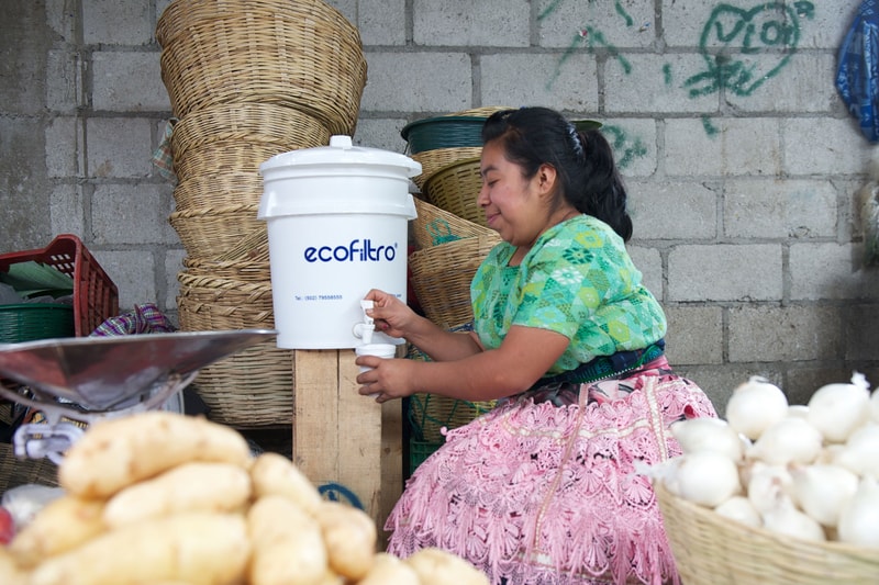 A woman in a green patterned blouse and pink lace skirt operates a white EcoFiltro water dispenser, surrounded by baskets and produce in an outdoor market.