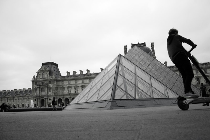 People skateboarding near the glass pyramid of the Louvre, with a stately historic building in the background and a wide, open plaza.