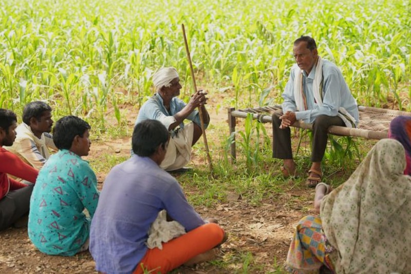 A group of rural villagers sits in a semicircle on the ground and a wooden bench in a field, listening to a man in traditional clothing speak.