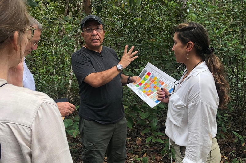 Several people stand in a leafy outdoor area as a man in a dark shirt and cap discusses a color chart held by a woman in a white blouse; others listen.