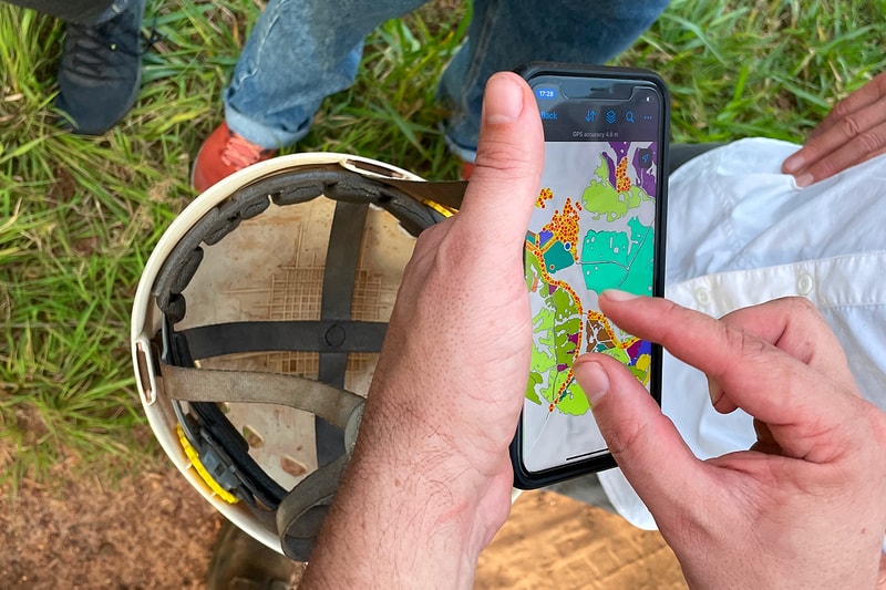 Person using a smartphone outdoors shows a colorful map on the screen while wearing a helmet nearby; grassy ground and a pair of boots visible in the background.