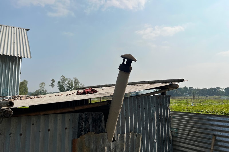 Rusted corrugated metal shed with a slanted roof, a vertical vent pipe pierced by a thick wooden beam, and a dusty rural landscape with fields in the distance.