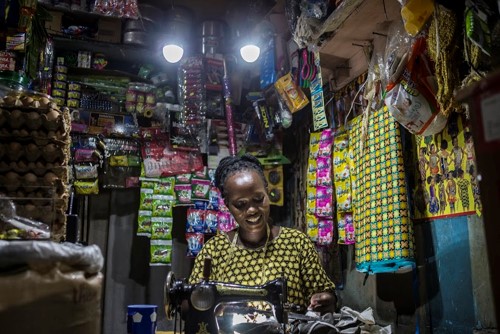 A smiling woman works at a crowded sewing counter inside a small shop, surrounded by colorful packaged goods and hanging trinkets, illuminated by overhead lights.