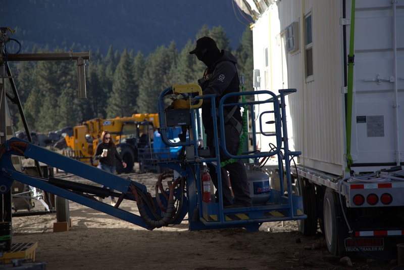Construction worker in dark clothing and hood on a blue aerial lift beside a white trailer, with machinery and trees in the background.