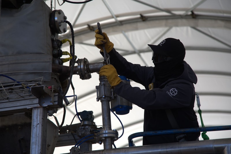 Worker in protective gear and yellow gloves operates industrial machinery on a scaffold, handling a wrench and cables inside a large sheltered structure.