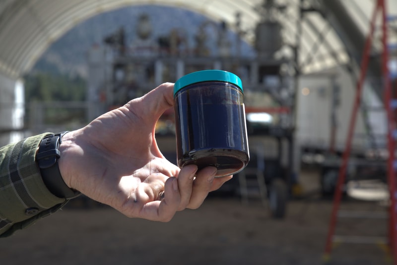 A hand holding a small dark blue jar with a teal lid in an outdoor industrial setting, blurred scaffolding and equipment in the background.