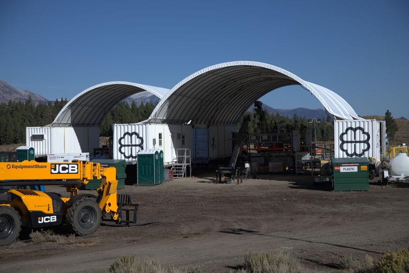 Temporary outdoor worksite with two white arched fabric structures, construction equipment (JCB) in yellow, portable toilets, portable offices, and containers against a mountainous backdrop.