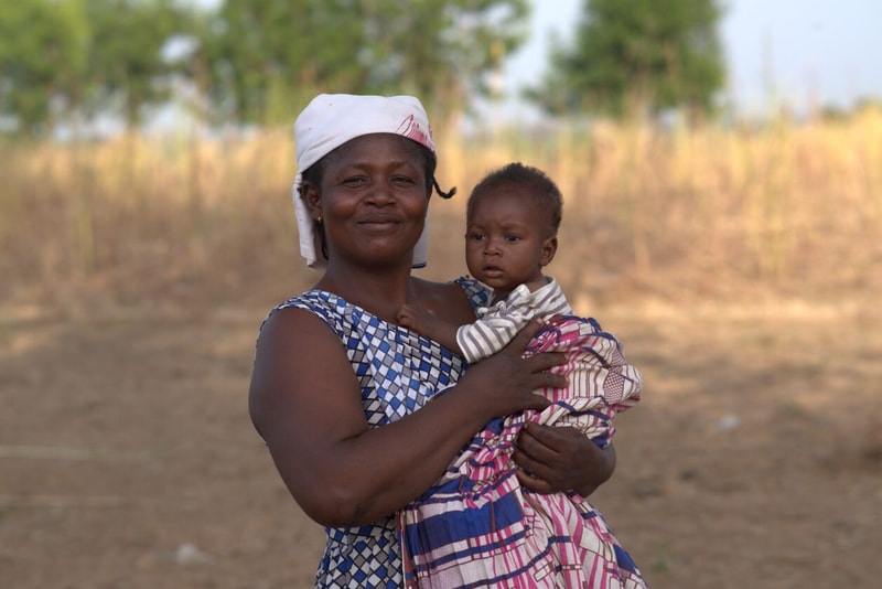 A smiling woman wearing a patterned dress and a white headscarf holds a small child wrapped in a colorful blanket outdoors in a dry field with trees in the background.
