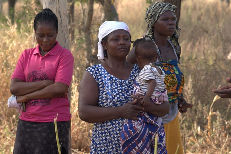Three African women stand outdoors in a dry field; one in a pink polo with arms crossed, a second woman holds a baby, and a third looks on, wearing colorful wrap.