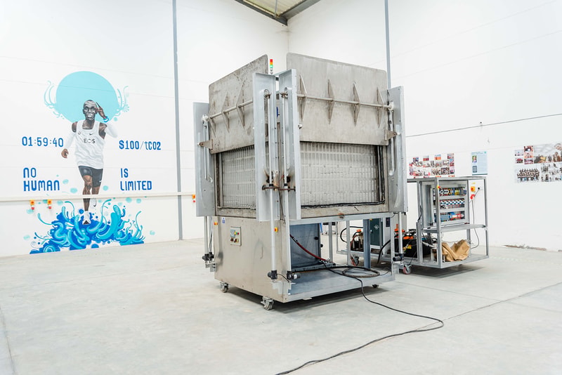 Industrial industrial-size machine in a bright room with white walls; large metal apparatus on wheels, cables on the floor, and a control console in the background.