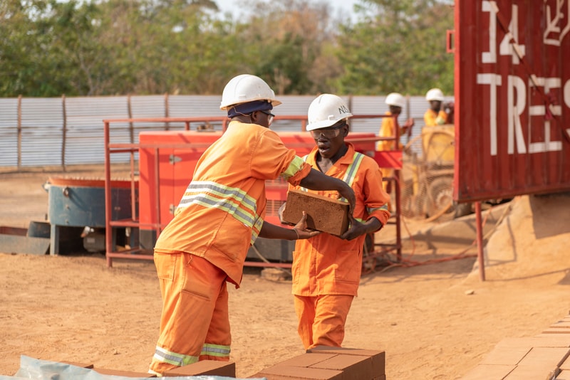 Two construction workers in orange uniforms and white hard hats pass a brick between them at a dusty outdoor site, with red equipment and a shipping container in the background.