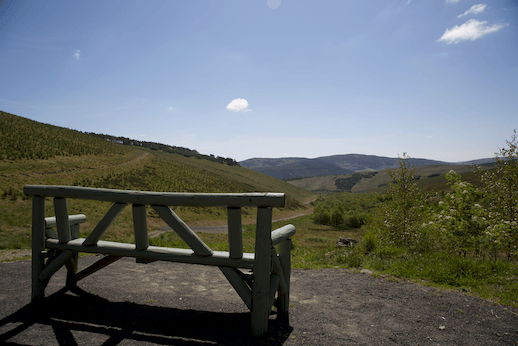 A rustic wooden bench on a gravel overlook facing rolling hills, green valleys, and a clear blue sky with a few scattered clouds.