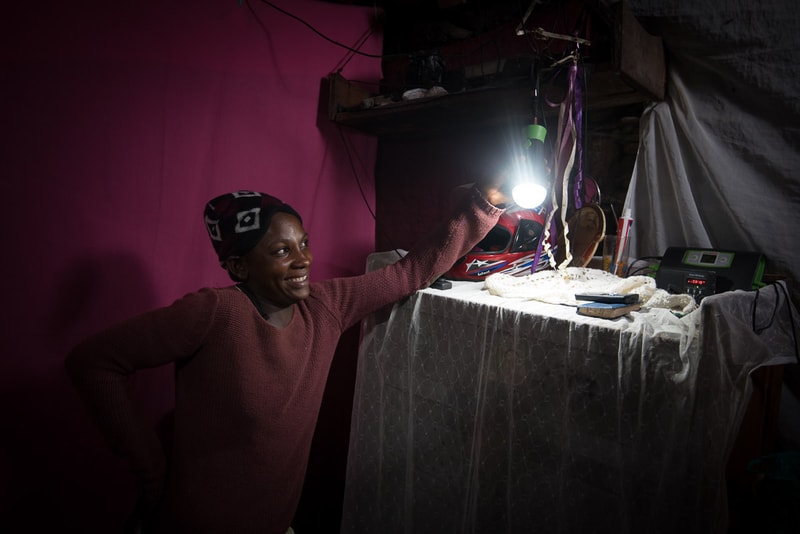 A person wearing a pink sweater and a patterned headwrap stands beside a table, smiling, with a bright work light and a hanging chain of decorations overhead in a dimly lit room.