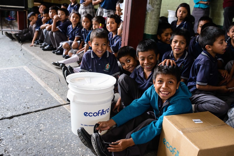Group of smiling schoolchildren in navy uniforms sit along a wall, with a boy in teal jacket at front and a white EcoFiltr bucket and cardboard box nearby.