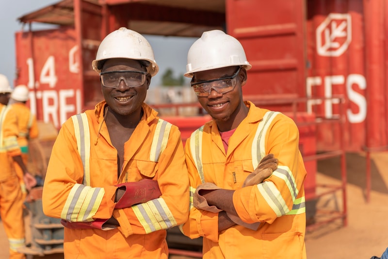 Two construction workers in orange safety suits and white hard hats smile with arms crossed, standing in front of red industrial equipment and containers.