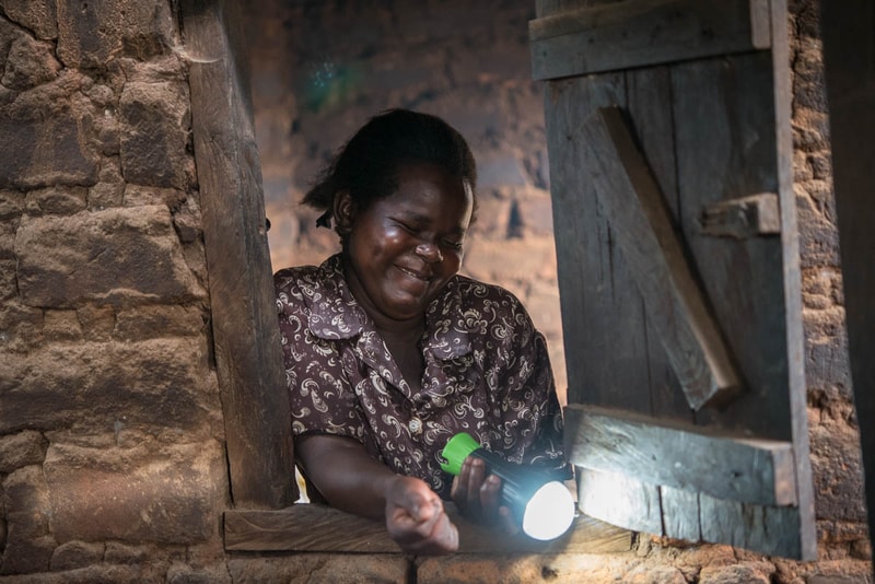 A smiling woman stands by a rustic brick and wood window, holding a lit flashlight as she leans on the sill, bathed in warm, soft light.