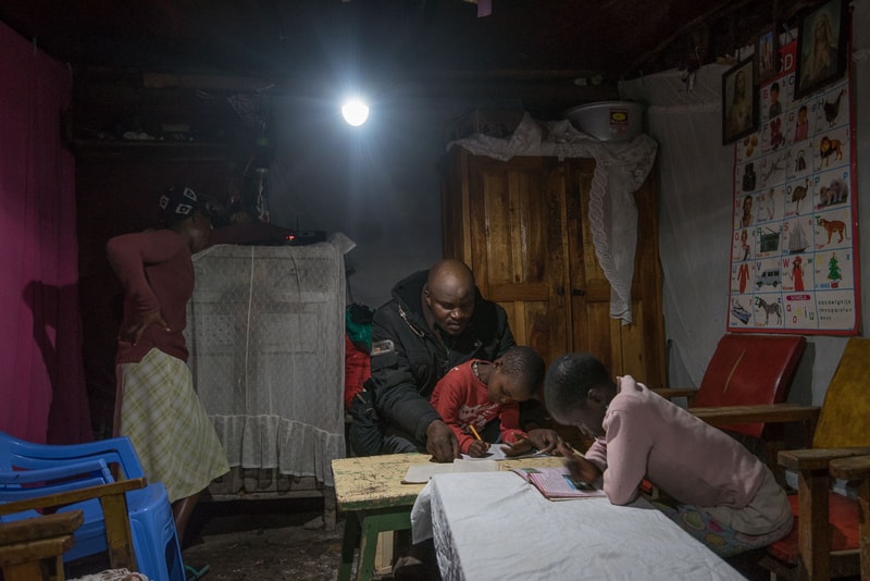 A man helps two children study at a small table in a dimly lit room; a bright bulb hangs overhead, with a wardrobe, chairs, and a colorful alphabet poster on the wall.