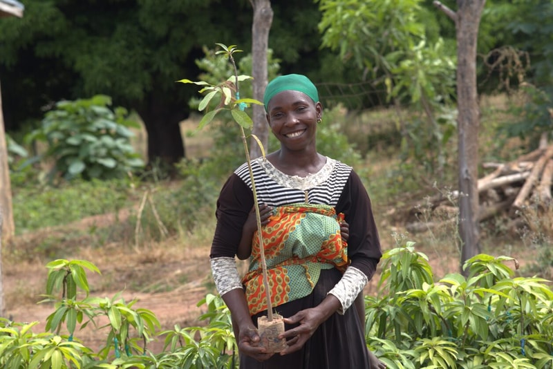 A smiling woman wearing a green headscarf and patterned fabric wrap holds a small potted sapling in a garden with green plants and trees in the background.