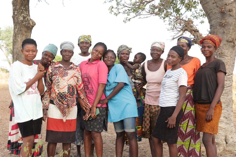 A group of diverse women and a child stand together outdoors under a tree, smiling and dressed in colorful clothing and headwraps.