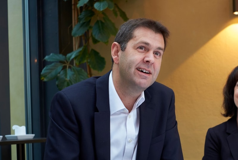 A man in a dark suit and white shirt sits indoors, looking slightly to the side with a smile, next to a woman, with a plant and warm wall lighting in the background.
