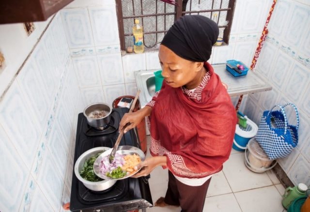 A woman wearing a red shawl and black headscarf cooks on a small gas stove, stirring vegetables in a pan while adding food to a plate in a compact kitchen.