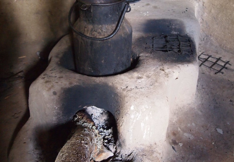 Charred, cracked stove with a soot-blackened kettle on a dusty, clay-like surface; a burnt log protrudes from a hollow opening, and metal grate lies nearby.