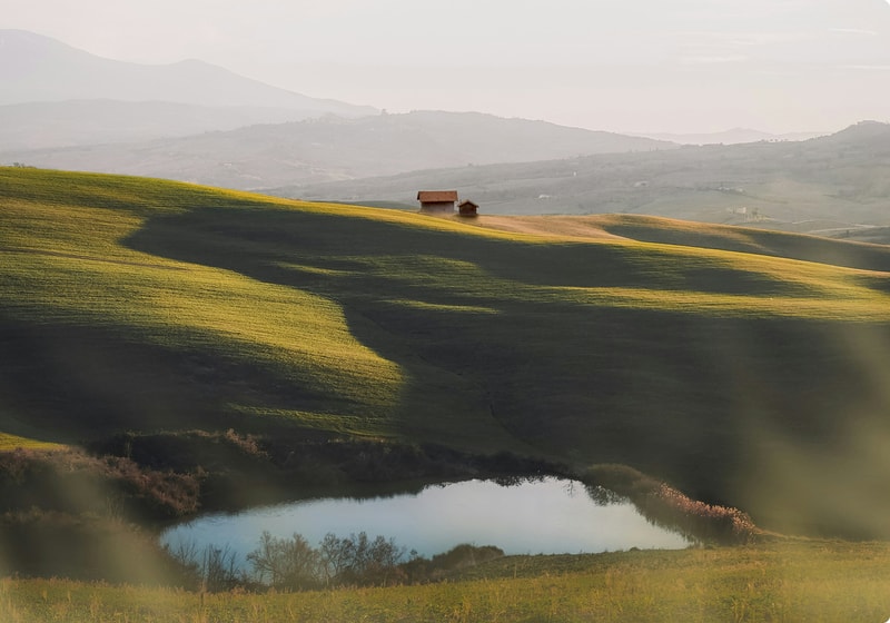 Rolled green hills at golden hour with two small wooden houses atop a ridge, a calm pond in the foreground, and distant hazy mountains on the horizon.