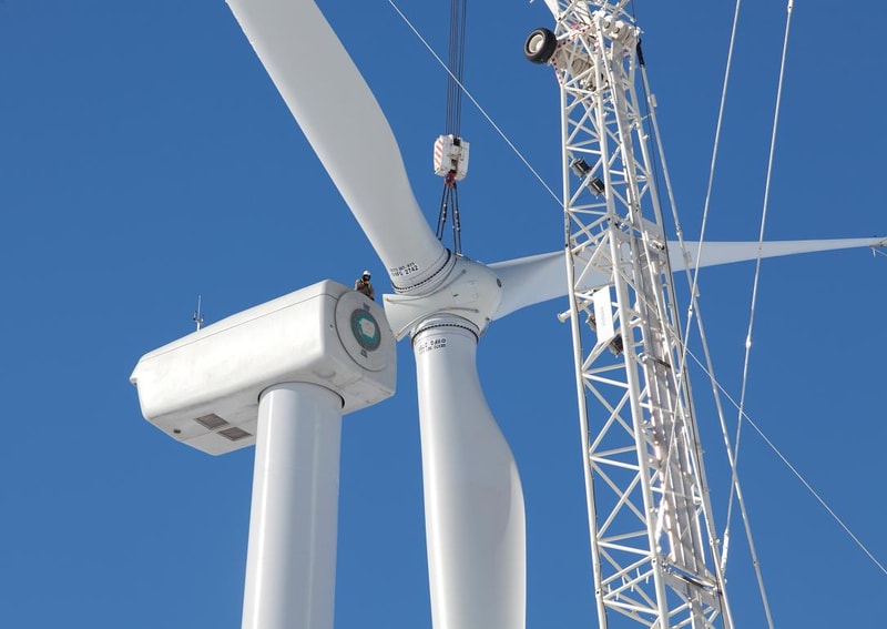 Wind turbine blades and nacelle under construction, workers on a machine hub, and a lattice crane against a clear blue sky.