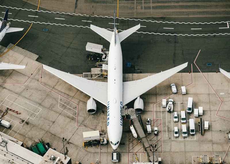 Aerial view of a large white United aircraft parked at a gate, connected to a jet bridge with ground crew vehicles, cargo carts, and service equipment around.