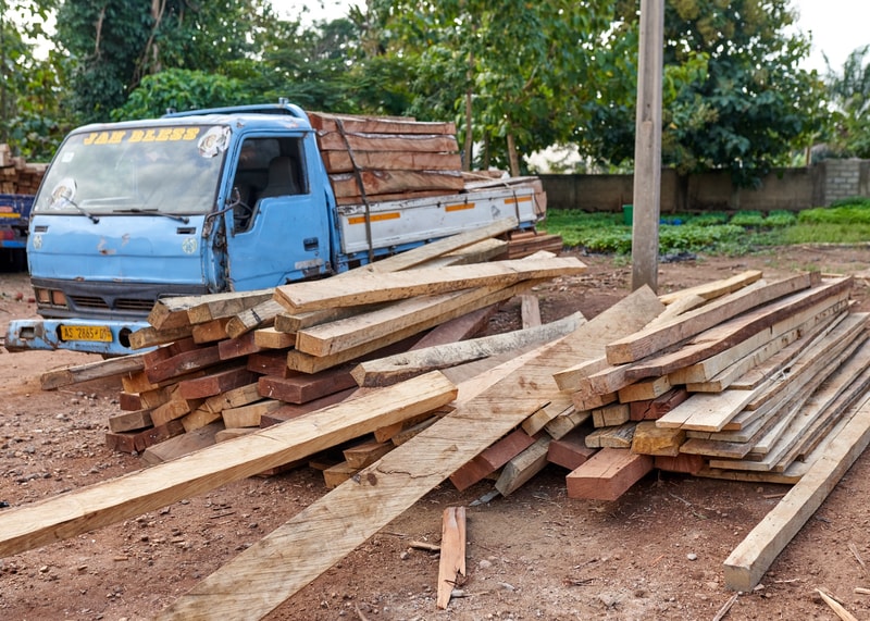 Pile of weathered wooden planks and beams stacked near a blue truck on a construction site, with dirt ground and green trees in the background.