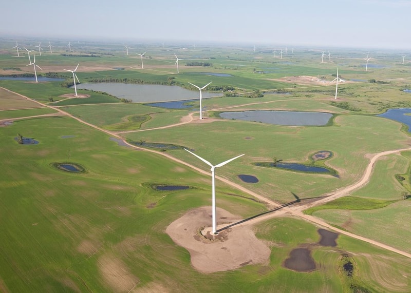Aerial view of a wind farm with white wind turbines scattered across green fields, dirt roads, and small ponds.
