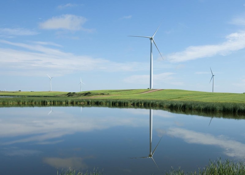 Wind turbines on a grassy hill beside a calm body of water, reflecting the turbines and blue sky with scattered clouds.