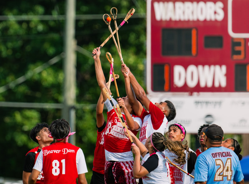 Lofters of a red-team water polo game raise sticks toward the sky as teammates in red and white shirts gather under a scoreboard on a sunny day.