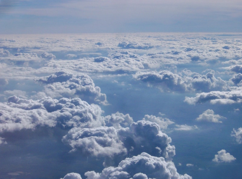 Aerial view of a vast sea of fluffy white clouds stretching to the horizon, with soft blue sky above and varied cloud formations casting gentle shadows.