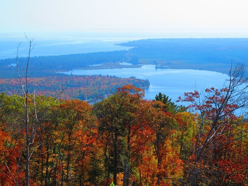 Vibrant autumn forest in foreground with red, orange, and yellow leaves; a blue lake and distant shoreline stretch across the mid and background under a clear sky.