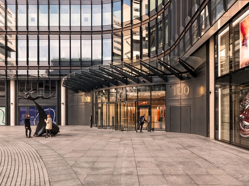Glass-fronted modern building entrance with a curved facade, large black metal awning, and the number 100 on doors; people and a dark sculpture in the plaza.