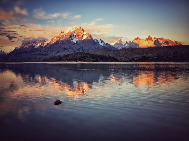 Sunlit snow-capped mountain range reflected in a calm lake at sunset, with warm orange-pink hues on peaks and a single rock in the water foreground.