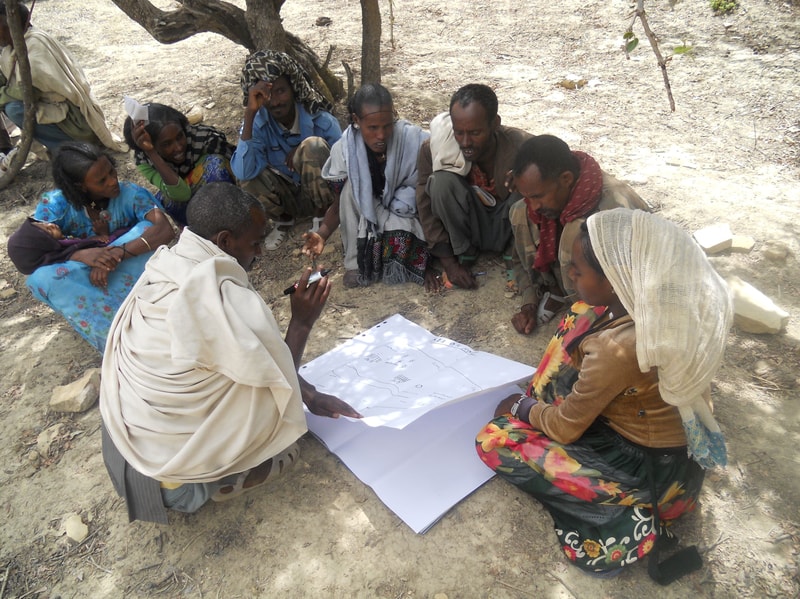 Group of people sitting on dusty ground in a circle outdoors, looking at a large sheet of paper with diagrams or drawings being discussed.