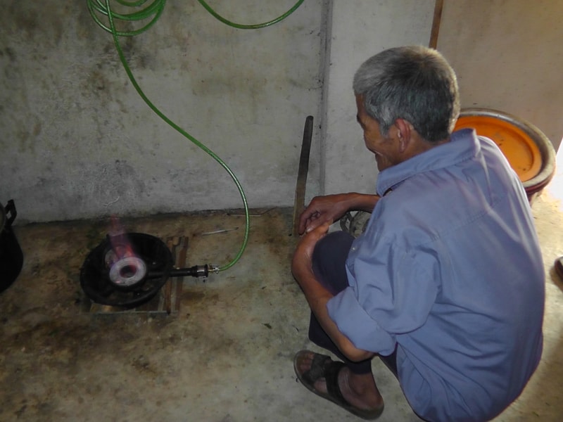 Man crouches on a concrete floor near a small repair setup, holding a tool with a green hose connected to a device emitting a small flame or heat.