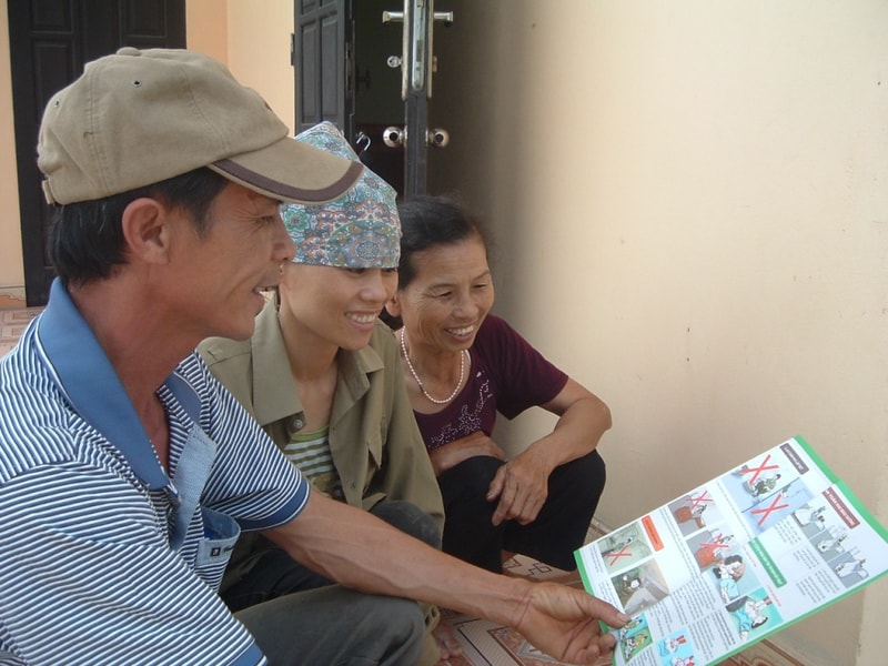 Three people sitting outside a building, a man in a cap pointing at a colorful instructional flyer with illustrations, as two women smile and look on.