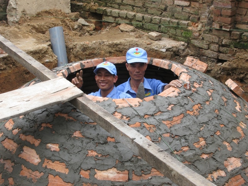 Two workers in blue uniforms and matching caps peek out from a circular brick and cement survival vault under construction, smiling and giving a peace sign.