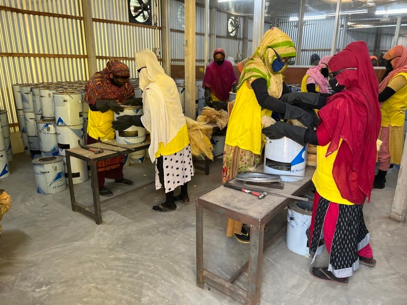 Women wearing colorful hijabs and yellow vests work at metal tables sorting and processing wool inside a well-lit industrial workshop with large barrels around.
