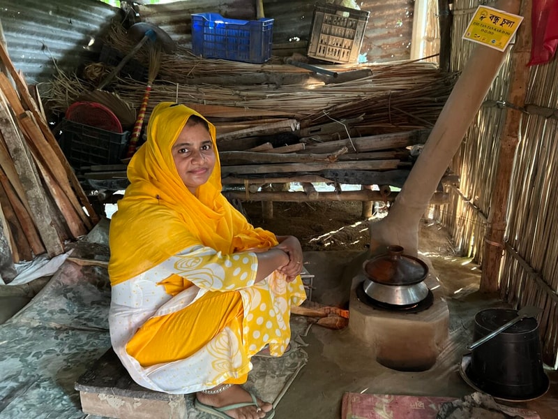 A woman in a bright yellow headscarf and dress sits inside a rustic, cluttered shed beside a clay stove with a metal pot, smiling at the camera.